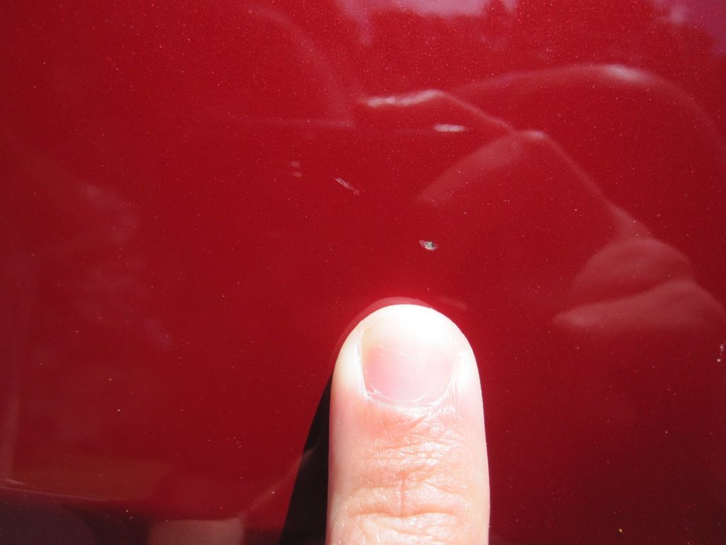 a very tiny white paint chip pointed out by a finger on a red vehicle